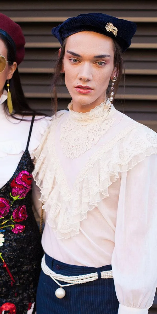 A person wearing a vintage-style blouse, pearl earrings, a brooch on a beret, and a pearl belt posing for a photo.