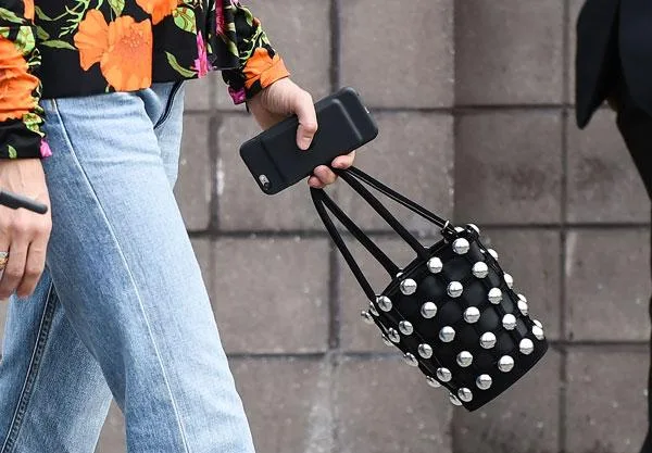 Person in jeans and floral top holding phone and studded black handbag.