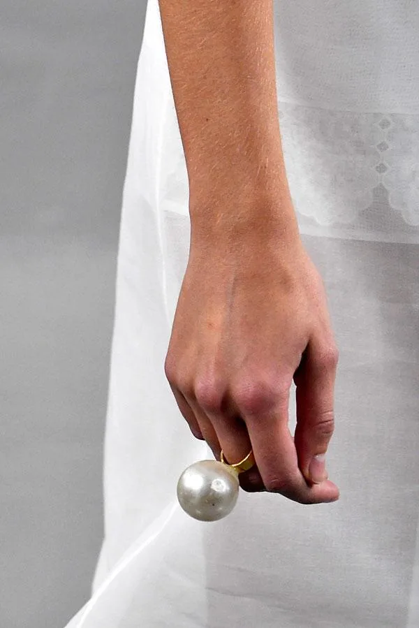 A hand holding a gold ring with a large pearl, against a white fabric background.