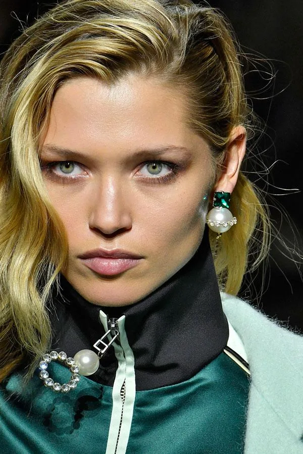 A model with blonde hair, green eyes, wearing pearl earrings and a high-collared green outfit on a fashion runway.