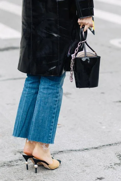 Black patent coat, cropped jeans, black handbag with chain, and black open-toed heels on urban street.