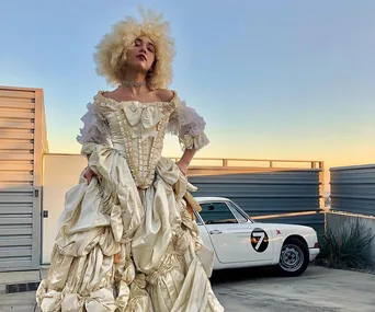 A person dressed as Marie Antoinette in a voluminous white gown, with a vintage car in the background.