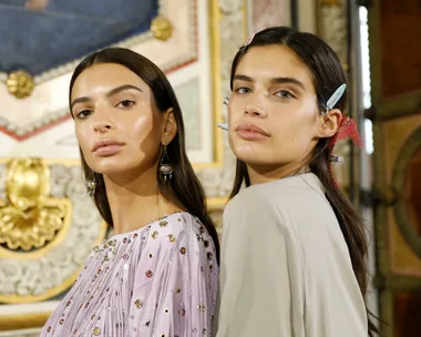 Two women with long dark hair, one wearing a beaded top, pose in an ornate room with hair clips and minimal makeup.