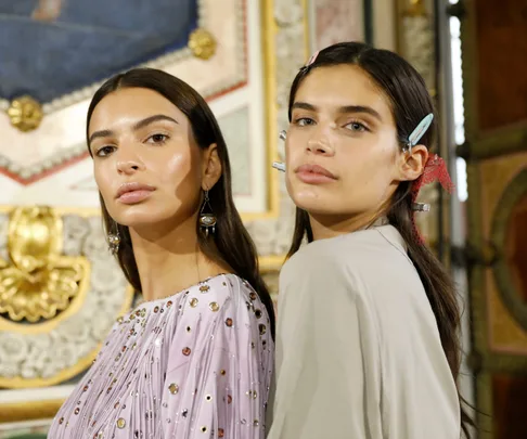 Two women with long dark hair, one wearing a beaded top, pose in an ornate room with hair clips and minimal makeup.