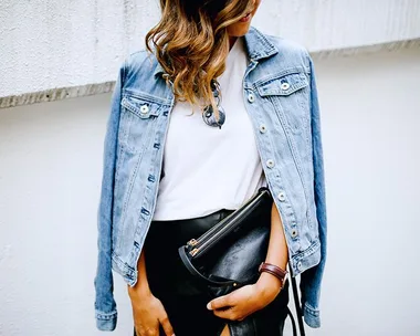 A person wearing a denim jacket over a white shirt, holding a black clutch bag.