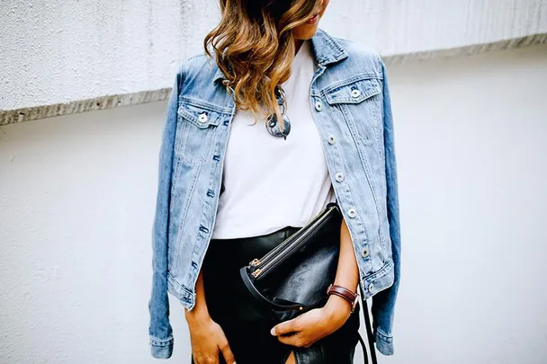 A person wearing a denim jacket over a white shirt, holding a black clutch bag.