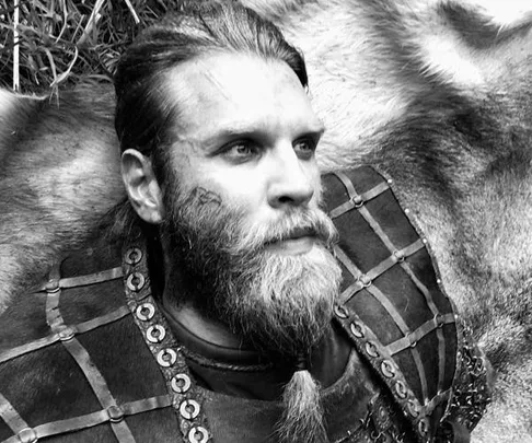 A grayscale image of a bearded man in medieval armor lying on the ground, looking upwards.