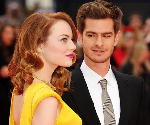 Emma Stone in a yellow dress and Andrew Garfield in a suit smiling at each other at a red carpet event.