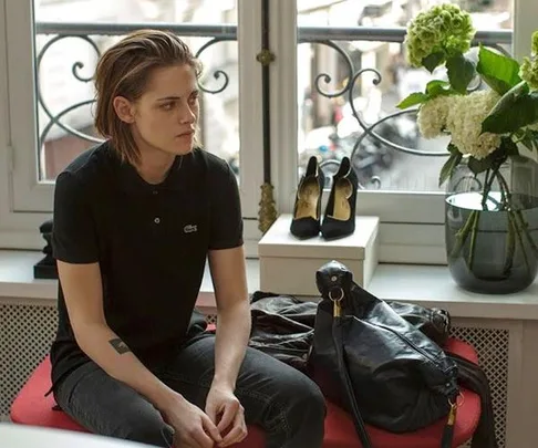 Woman sitting on sofa with leather handbag, black stiletto shoes on stand, flowers on window sill, looks melancholic.