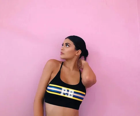 A woman in a sports bra stands against a pink wall, looking to her right with her left hand touching her hair.