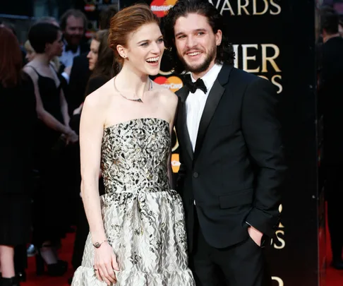 A smiling woman in a strapless dress stands next to a man in a tuxedo on a red carpet event.