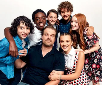 Stranger Things cast members grouped, smiling together, with an adult sitting in the center and kids surrounding him.