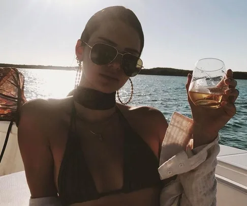 Woman in sunglasses and bikini holding a drink, sitting on a boat with a sunny lake and horizon in the background.