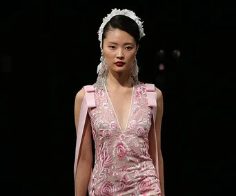 Model in a floral pink bridal gown with white headpiece and long earrings on runway, dark background.