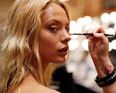 A woman with long blonde hair gets makeup applied with a brush in a well-lit setting.