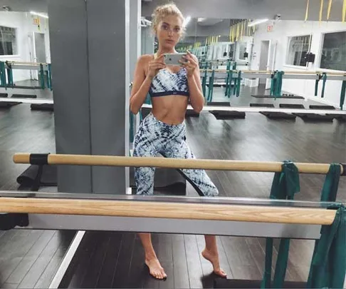 Young woman in a fitness studio taking a selfie, wearing a blue and white workout outfit by a ballet barre.