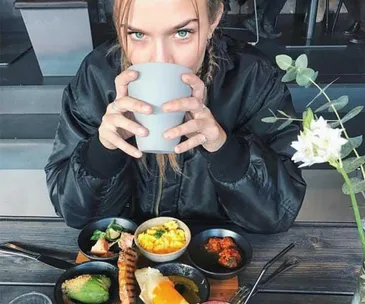 A person with a cup, sitting at a table with an assorted, healthy meal in small bowls and plates, wearing a black jacket.