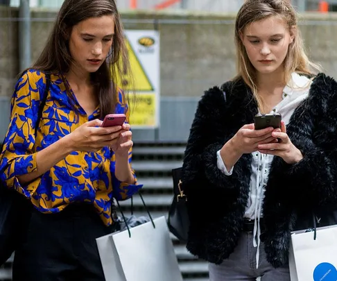 Two women standing, looking at their smartphones, holding shopping bags outside.