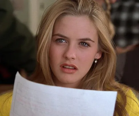 Alyssa Silverstone as Cher Horowitz, confused, holding a paper in a classroom scene from the movie "Clueless."