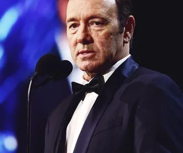 Kevin Spacey, wearing a black suit and bow tie, stands at a microphone against a blurred background at an event.