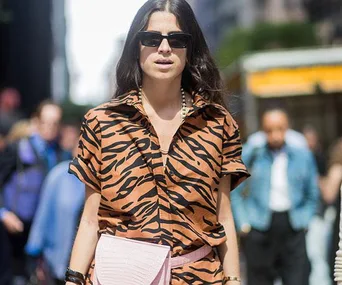 A woman in a tiger print shirt, sunglasses, and a pink bag walks down a city street during Fashion Month SS18.