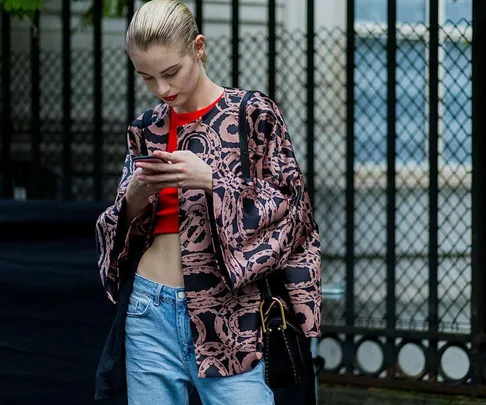 A woman in a patterned jacket and jeans looks at her phone while standing in front of a fence.