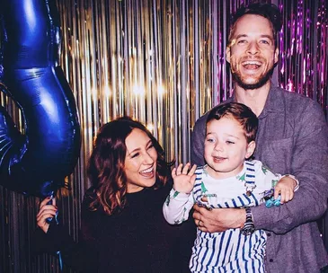 A smiling woman, man, and child in front of a metallic streamers backdrop, with the child holding a large blue number 3 balloon.