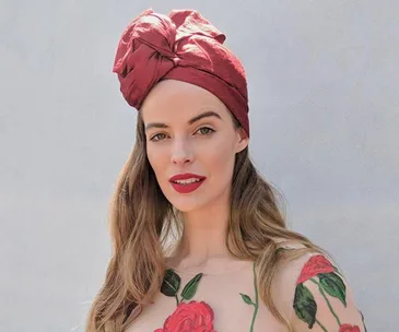 Woman wearing red head wrap and floral dress, posing against a light grey background.