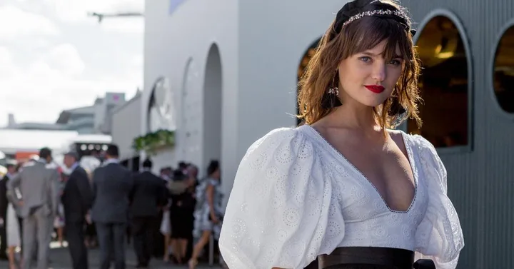 Woman in a white dress and headpiece at Derby Day 2017, with people and building in background. Bright, sunny day.