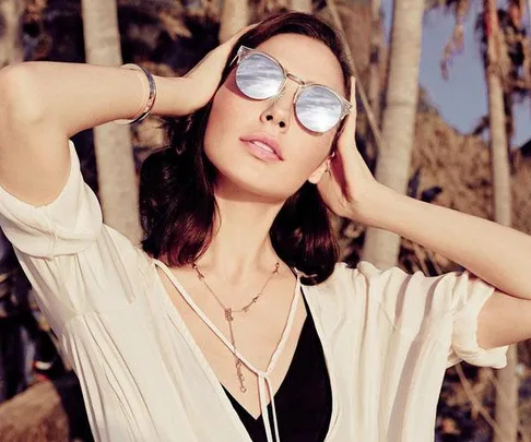 Gal Gadot, the actress of Wonder Woman, posing outdoors in reflective sunglasses, a white top, and jewelry, with her hands behind her head.