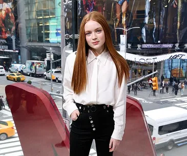 Sadie Sink in a white blouse and black pants stands with a city street visible through the window behind.