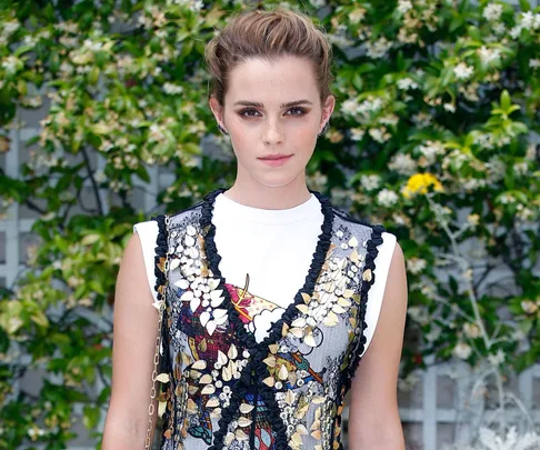 Emma Watson at an event, wearing a floral dress, standing in front of a foliage background.