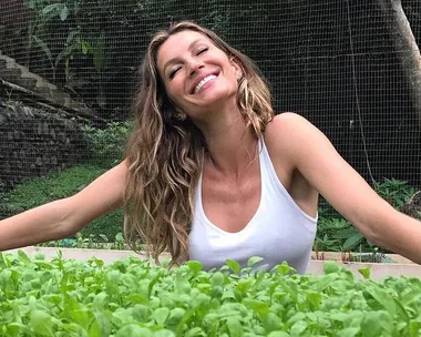 Woman with wavy blonde hair in a white tank top smiling and stretching arms out in garden, surrounded by green plants.