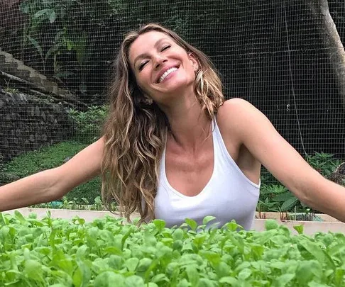 Woman with wavy blonde hair in a white tank top smiling and stretching arms out in garden, surrounded by green plants.