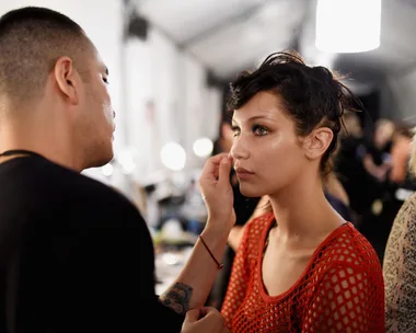 A makeup artist applying makeup to a person with dewy skin in a red top, in a well-lit room.