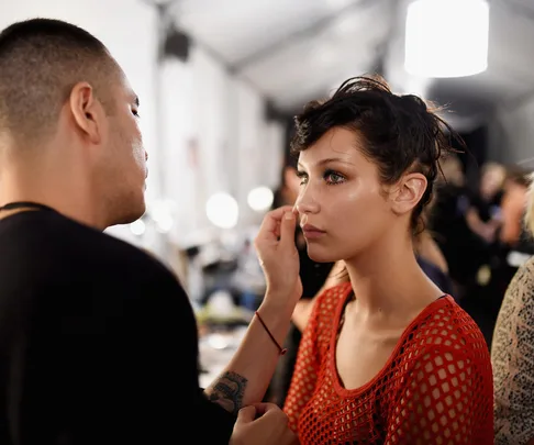 A makeup artist applying makeup to a person with dewy skin in a red top, in a well-lit room.