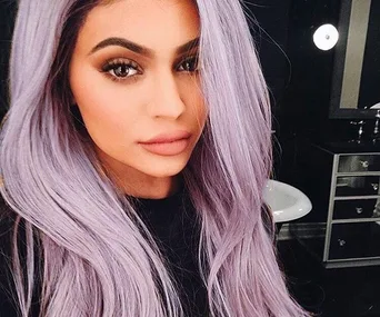 A person with long lavender hair and natural makeup, looking directly at the camera in a dimly lit room with a round mirror in the background.