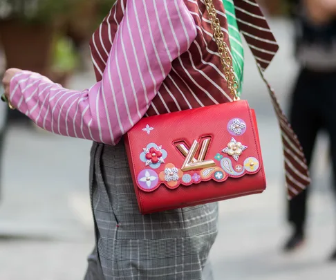 A person wearing a pink striped shirt and gray pants carries a red Louis Vuitton bag decorated with colorful floral patterns.