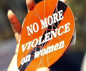 A person holds a circular sign reading "NO MORE VIOLENCE on women" in bold white letters on a red background.