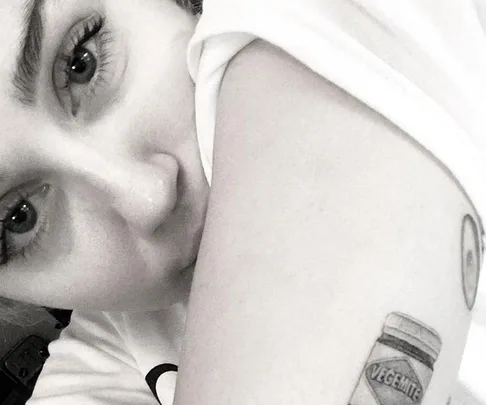 Close-up of a person's face resting on their arm, highlighting a tattoo of a Vegemite jar on the forearm.