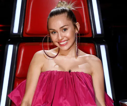 Miley Cyrus smiling and wearing a pink dress while sitting in a red chair from "The Voice."