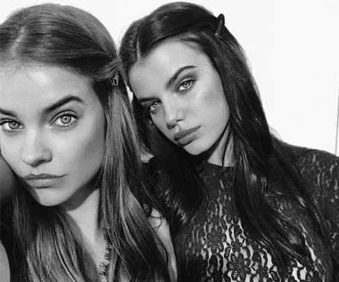 Two women with long hair and strong makeup, one with hair clips, pose together looking serious in black and white.