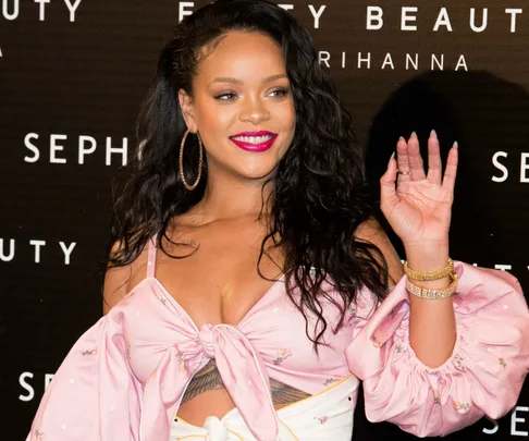 A woman in a pink top with a bow and hoop earrings, smiling and waving at a Sephora event. Background says "Fenty Beauty" and "Sephora".