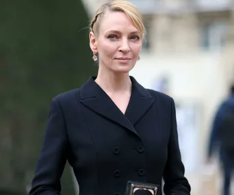 A woman with blonde hair styled in braids, wearing a black double-breasted coat and pink earrings, standing outdoors.