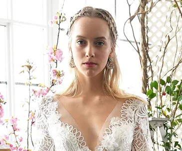 Bride in a white lace wedding dress with braided hair, standing in front of flowering pink branches and greenery.