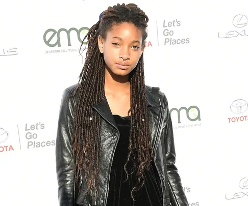 Person with long braids in black leather jacket at an Environmental Media Association event backdrop.