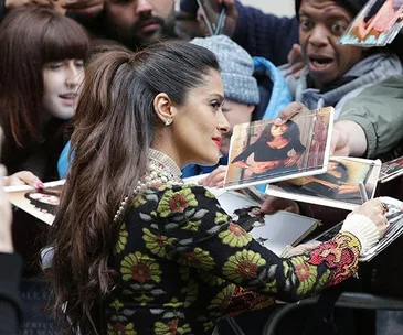 Actress signing autographs for fans, smiling with hair in ponytail, wearing floral top, surrounded by exuberant crowd.