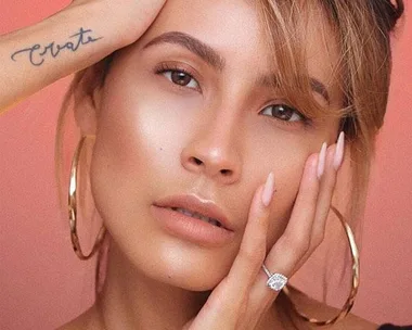 A woman with smooth, glowing skin, wearing hoop earrings and a diamond ring, with a tattoo on her arm that reads "Create."