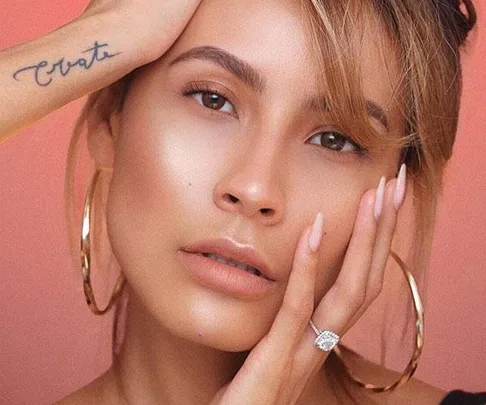 A woman with smooth, glowing skin, wearing hoop earrings and a diamond ring, with a tattoo on her arm that reads "Create."