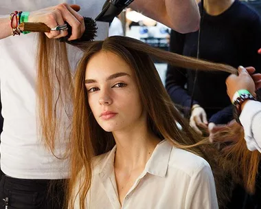 A model with long brown hair receiving a blowout at a salon, with stylists using brushes and a hairdryer.